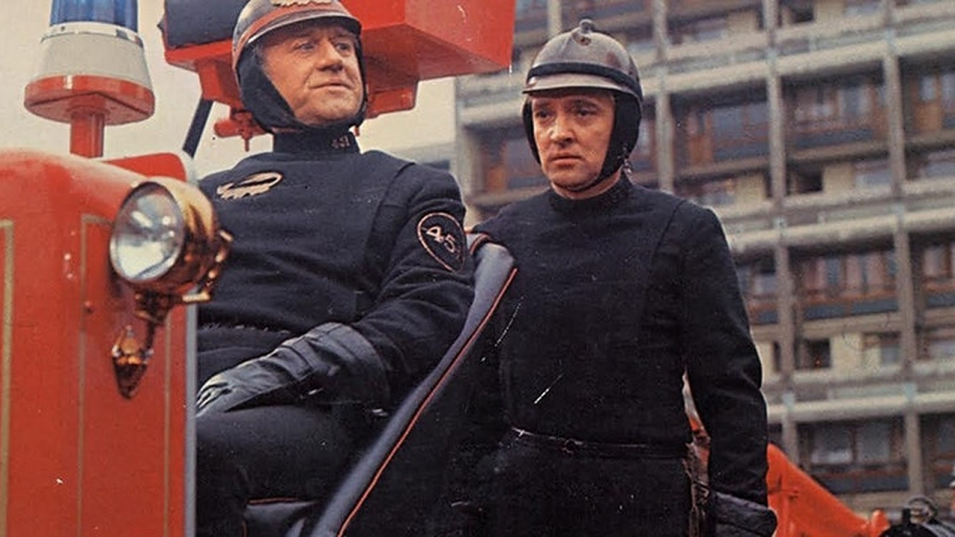Film still from .Fahrenheit 451'. Two men in firefighter costumes and helmets standing on a fire truck.