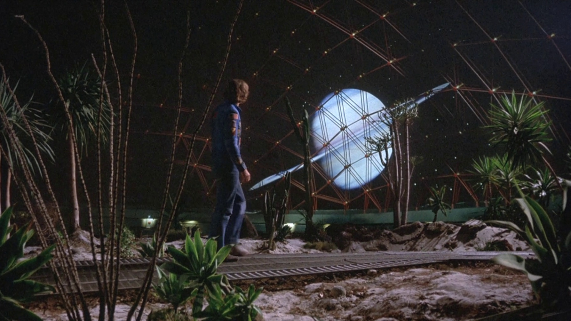 Film still from 'Silent Running'. Man in astronaut suit in a space dome, surrounded by plants staring out at a planet.