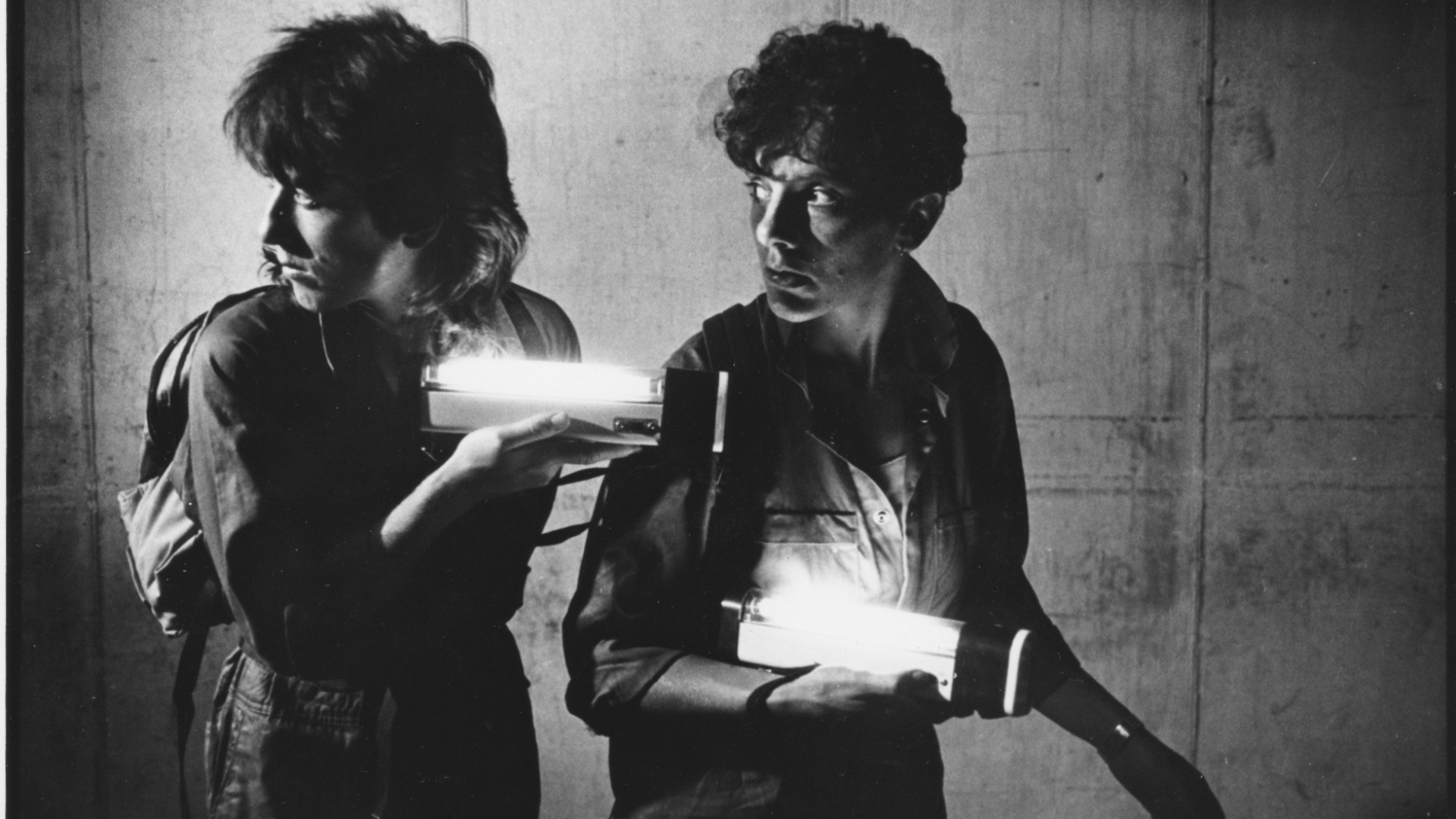 Film still from 'On Guard'. In black and white, two people hold flashlights and walk through a cement corridor.