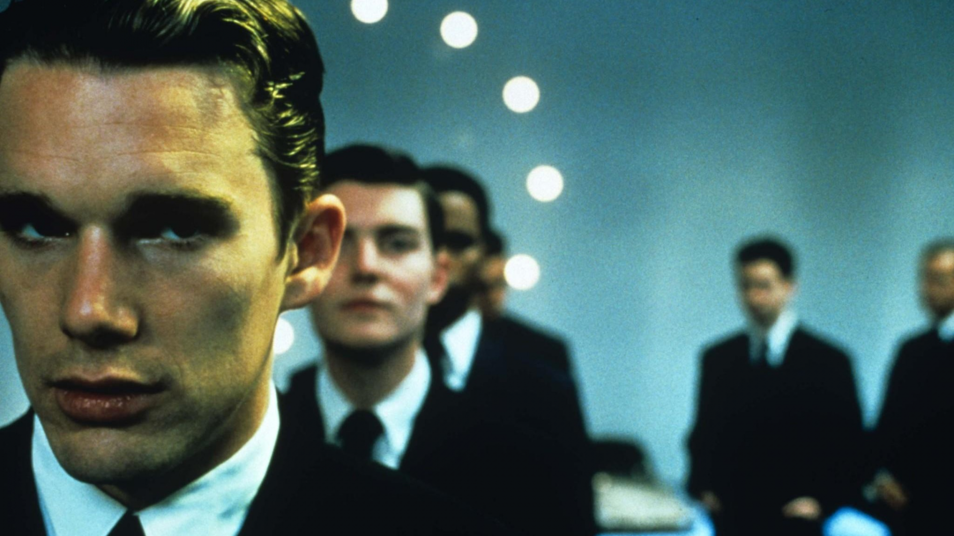 Film still from 'Gattaca'. Men dressed in suits in a stark room, one in the foreground with an ominous expression on his face.