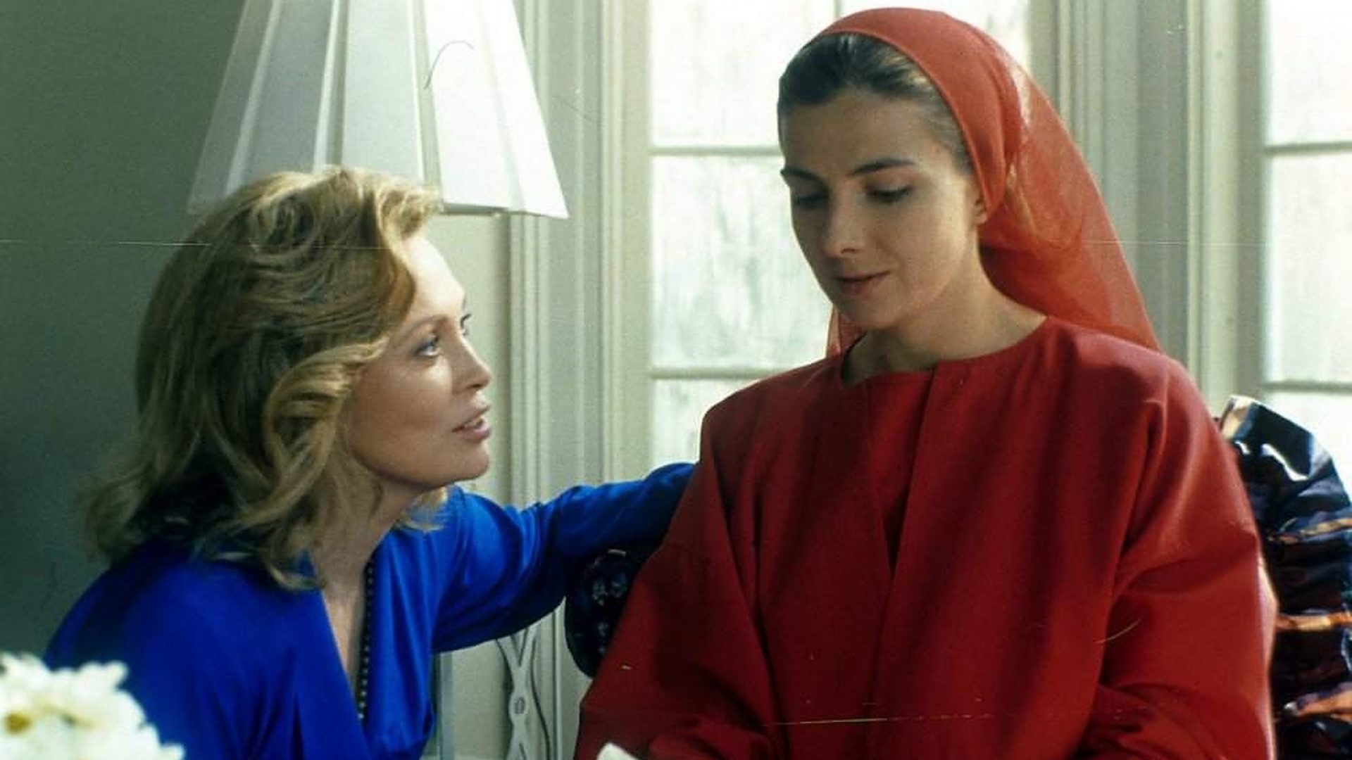 Film still from The Handmaid's Tale. Two women sit down together talking, one dresses in blue the other dressed in red.