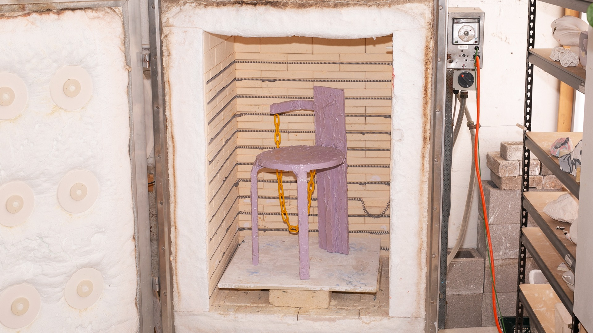 Photo of a kiln with an open door, inside the kiln is a lilac chair with a decorative orange chain.