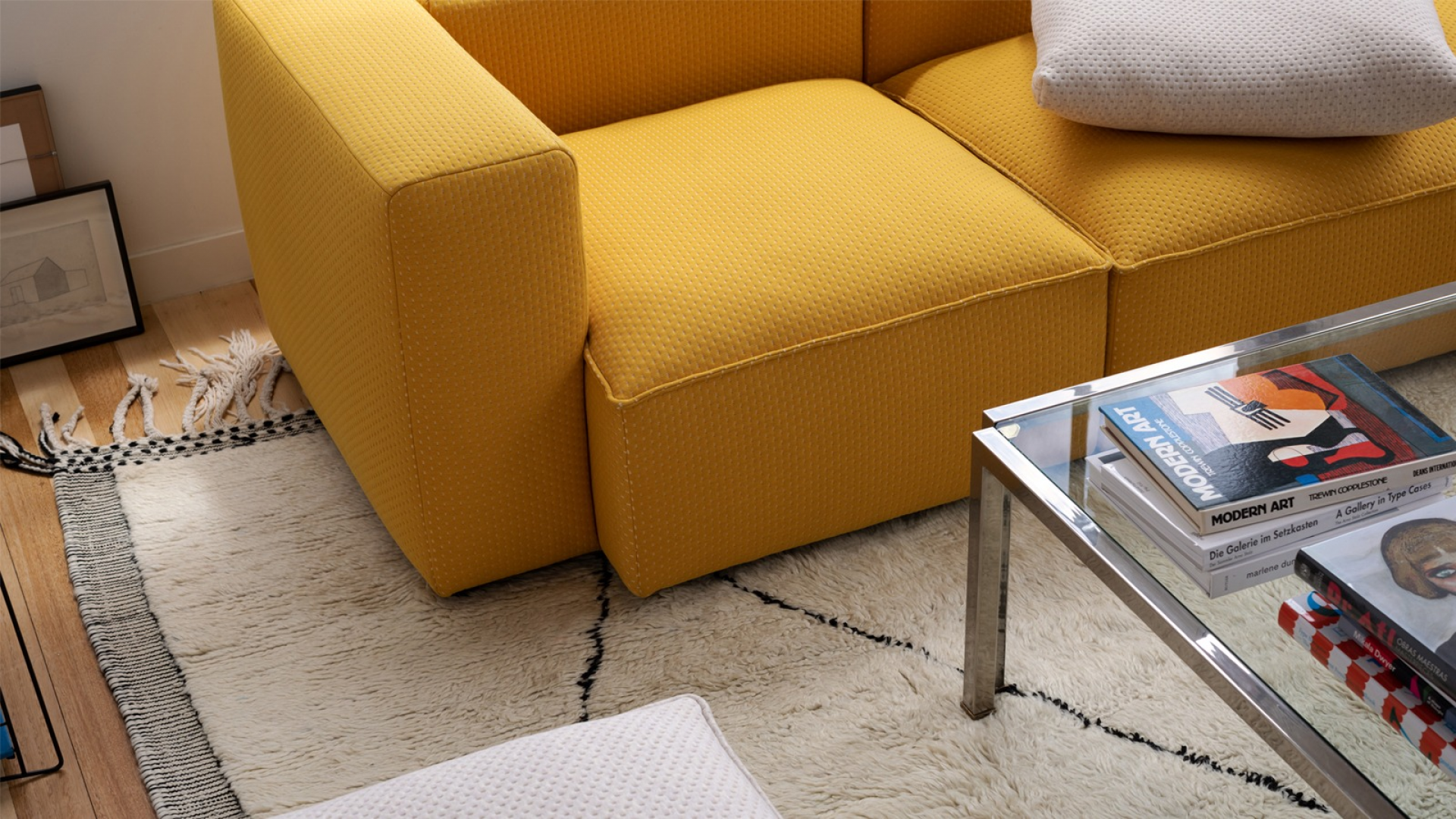 Photo taken from above of a boxy yellow sofa on a shaggy natural wool rug, situated in a living room style space with a coffee table with art books.