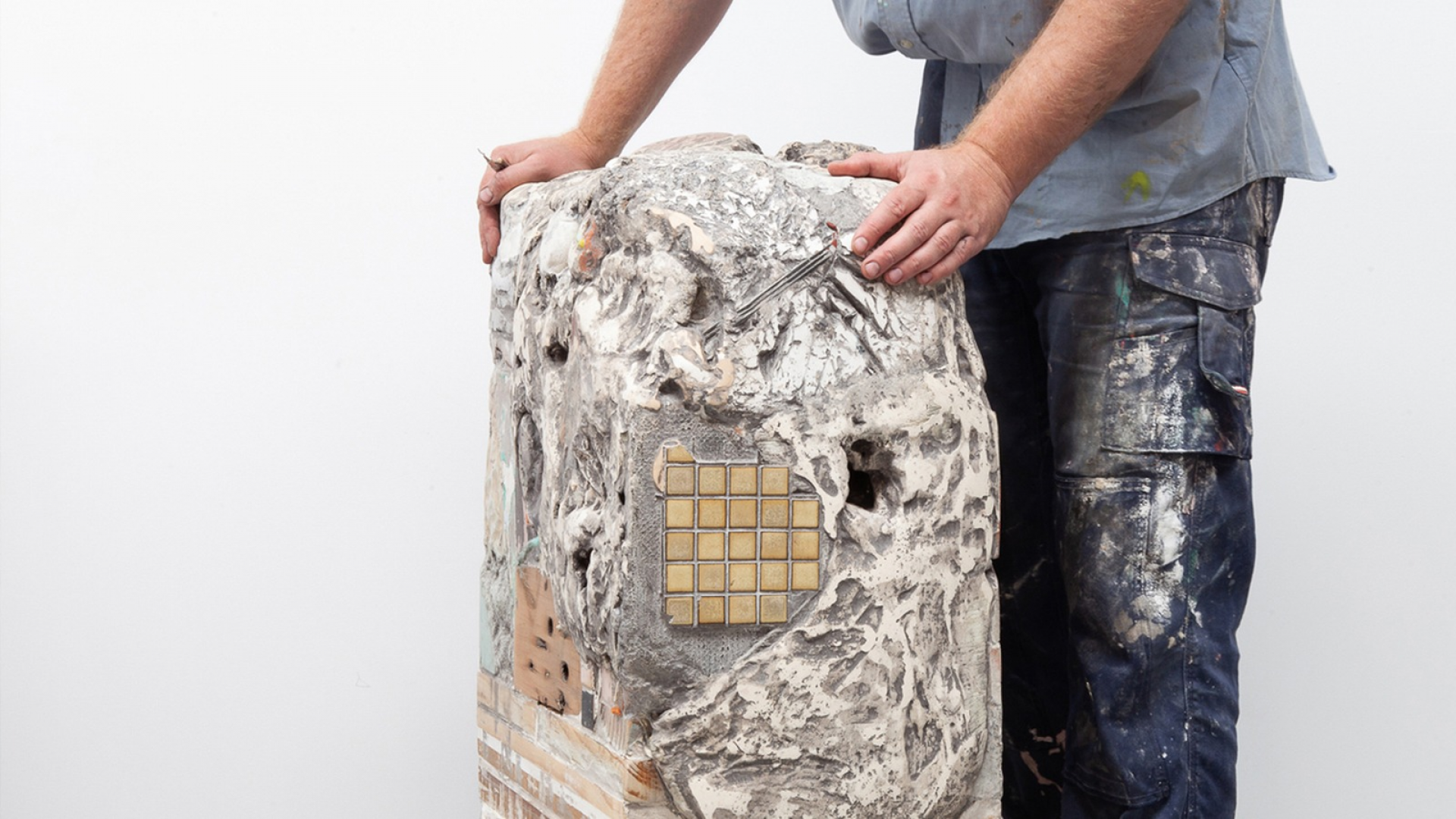 Photo of a person standing beside, and gripping with two hands, a waist-height square-column-shaped sculpture made from plaster, concrete, aerated concrete, concrete sheet, rocks, tiles, oregon, American oak, MDF, ply, various foams, string, wire, canvas, acrylic, Apoxie Sculpt, putty, wax, glue, silicon, rubber with varied surface texture, muted colours and a white-wash effect finish.