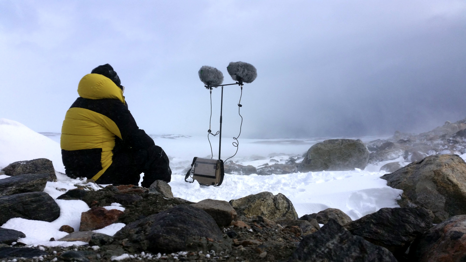A man in a yellow and black puffy jacket sits to the left on 2 fluffy microphones on tall stands. He is in the now in Antarctica
