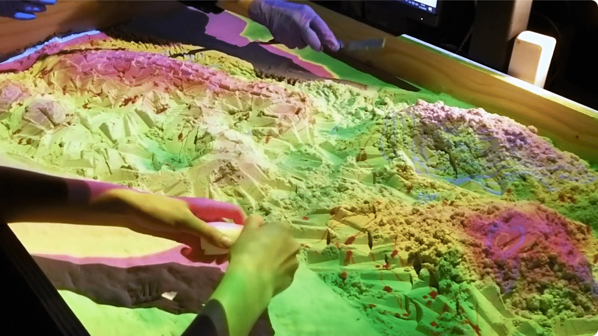 A pair of hands use a tool to shape a mass of sand in a table. A range of bright color gradients are projected atop the sand.