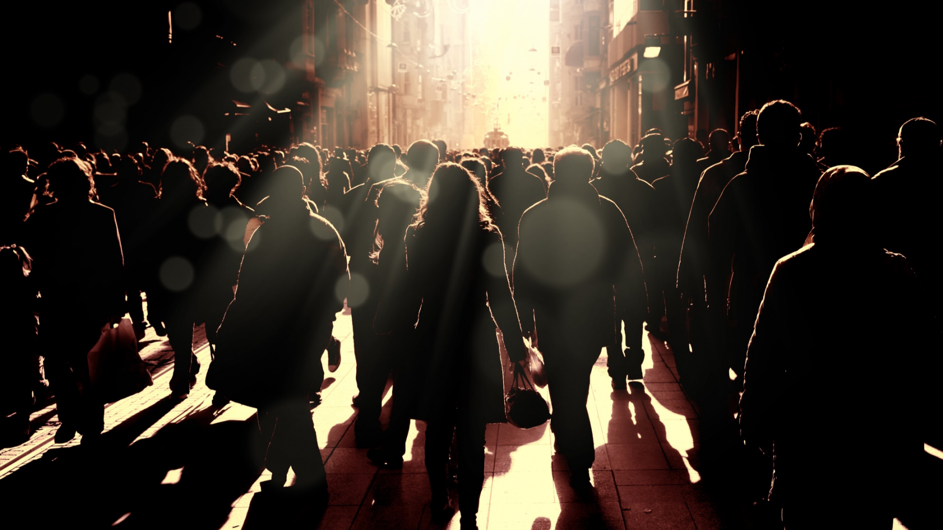 A crowd standing in a city street with light shining down on them, which also casts shadows across the crowd