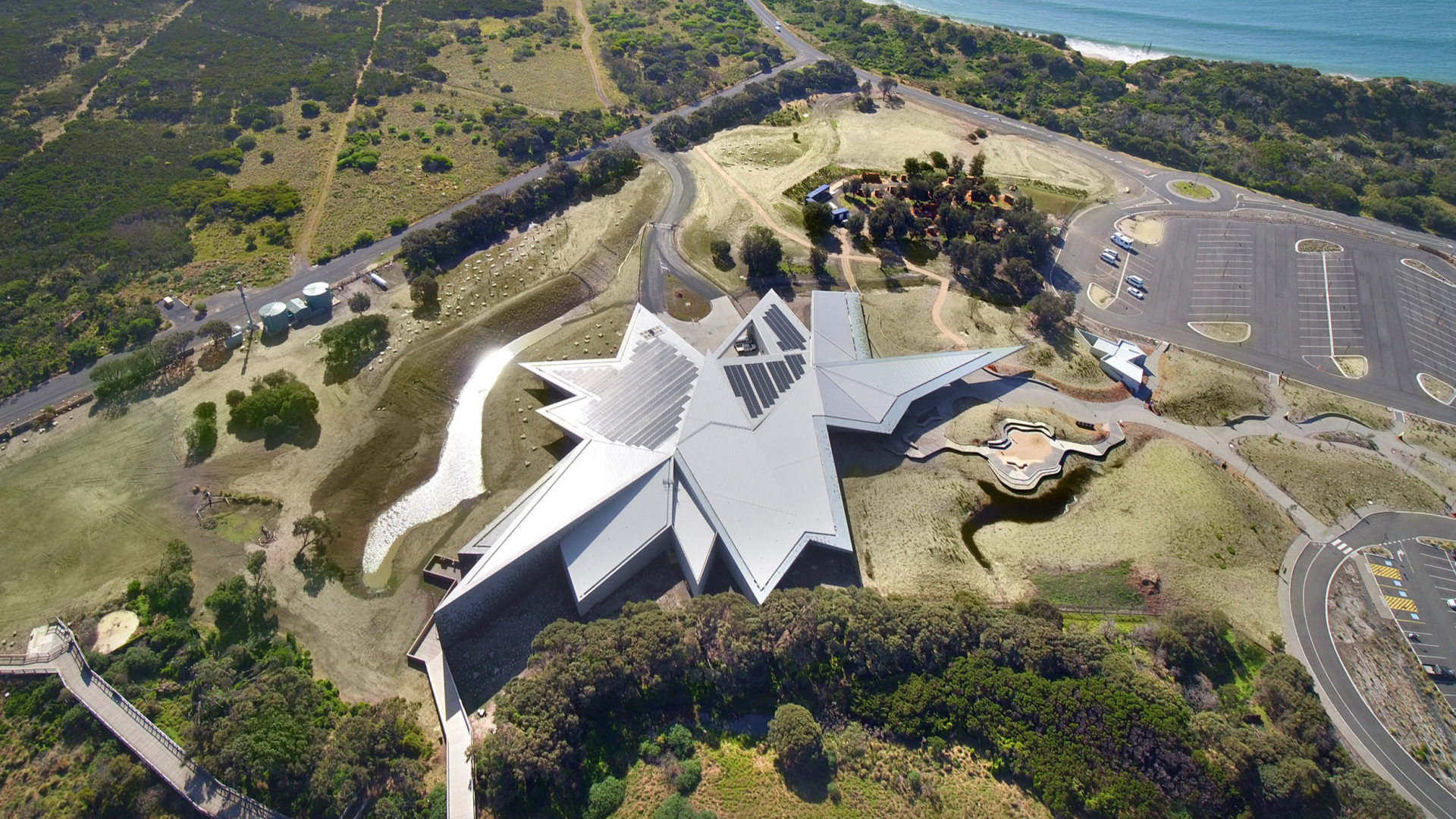 Aerial photograph of the Philip Island Nature Parks Visitors Centre. An angular silver building in a green landscape by the beach.