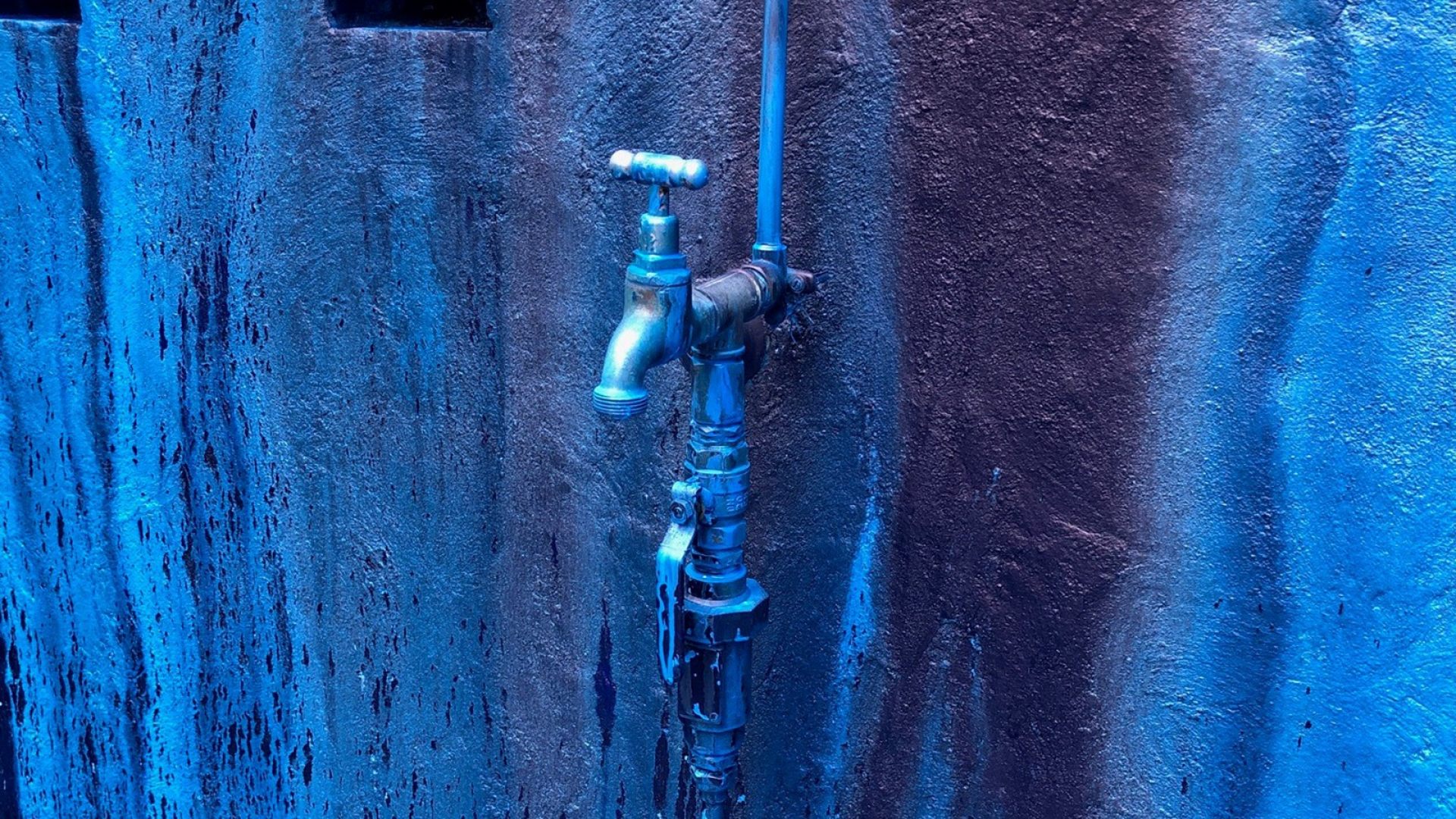 A silver tap is mounted to a wall. The whole image has a blue wash